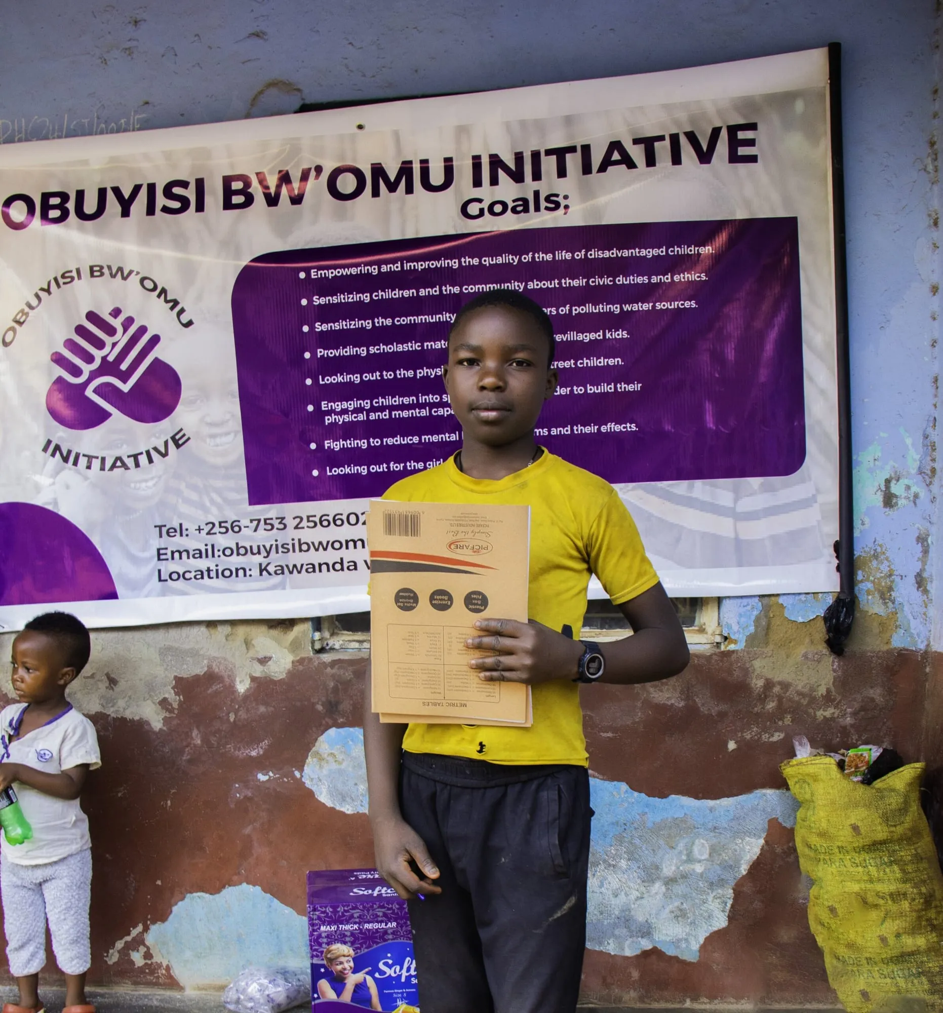 A student holding new exercise books in front of the Obuyisi bw'Omu Initiative banner
